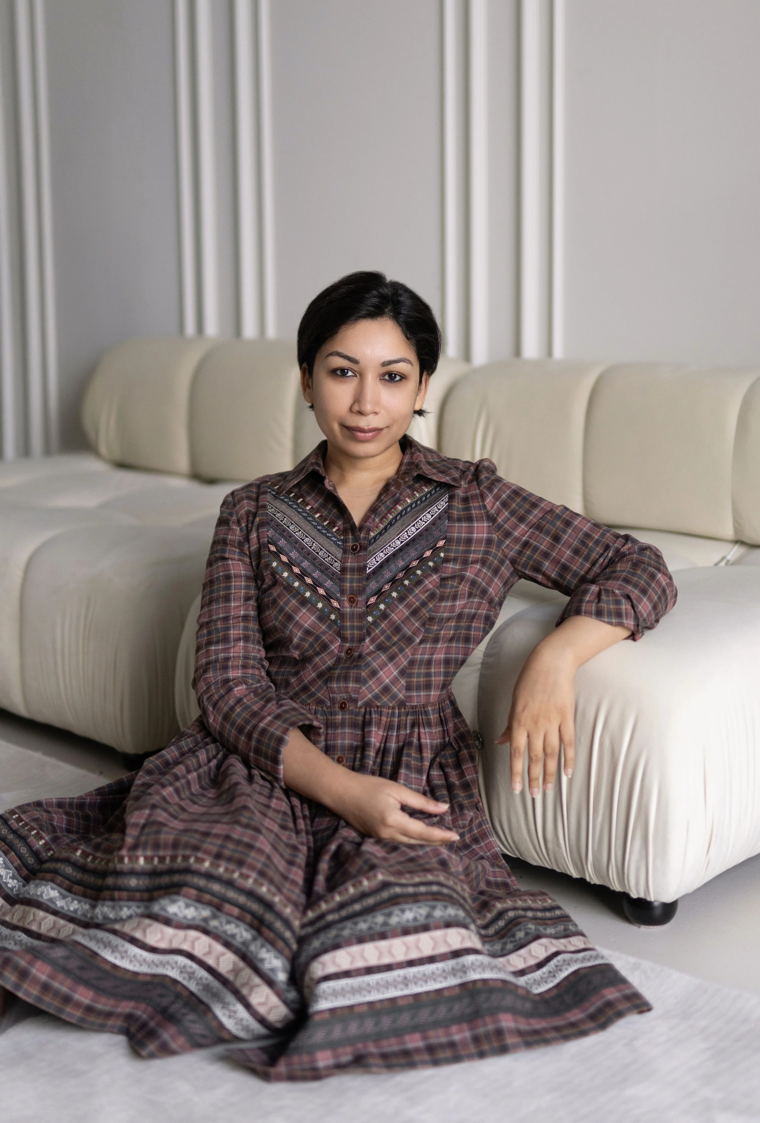 Arya Rose, trainee in clinical psychology. A woman with short black hair sitting on the floor in front of a light-colored sofa, wearing a plaid dress with embroidery on the chest.