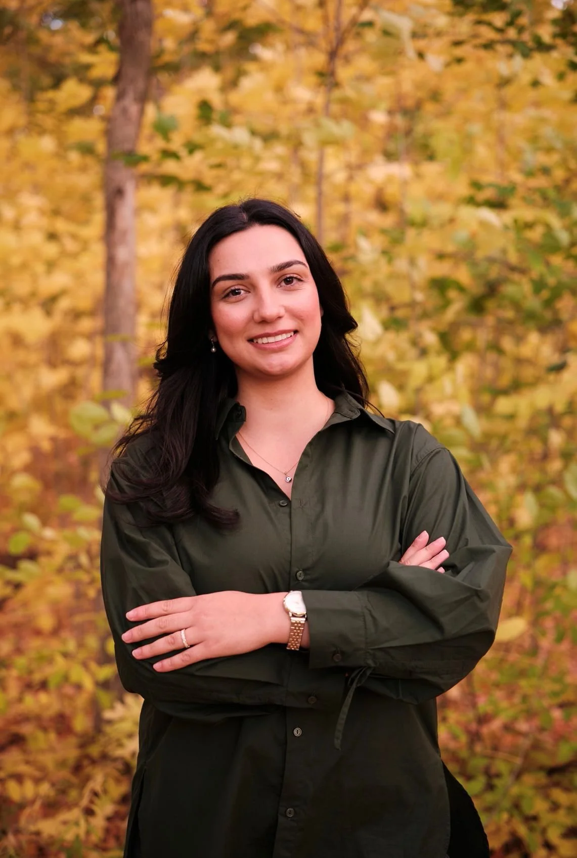 Mariana Profka, Registered Psychotherapist (Qualifying). A young woman with dark hair and light skin smiling with her arms crossed, standing outdoors in a forest with yellow and green autumn leaves.