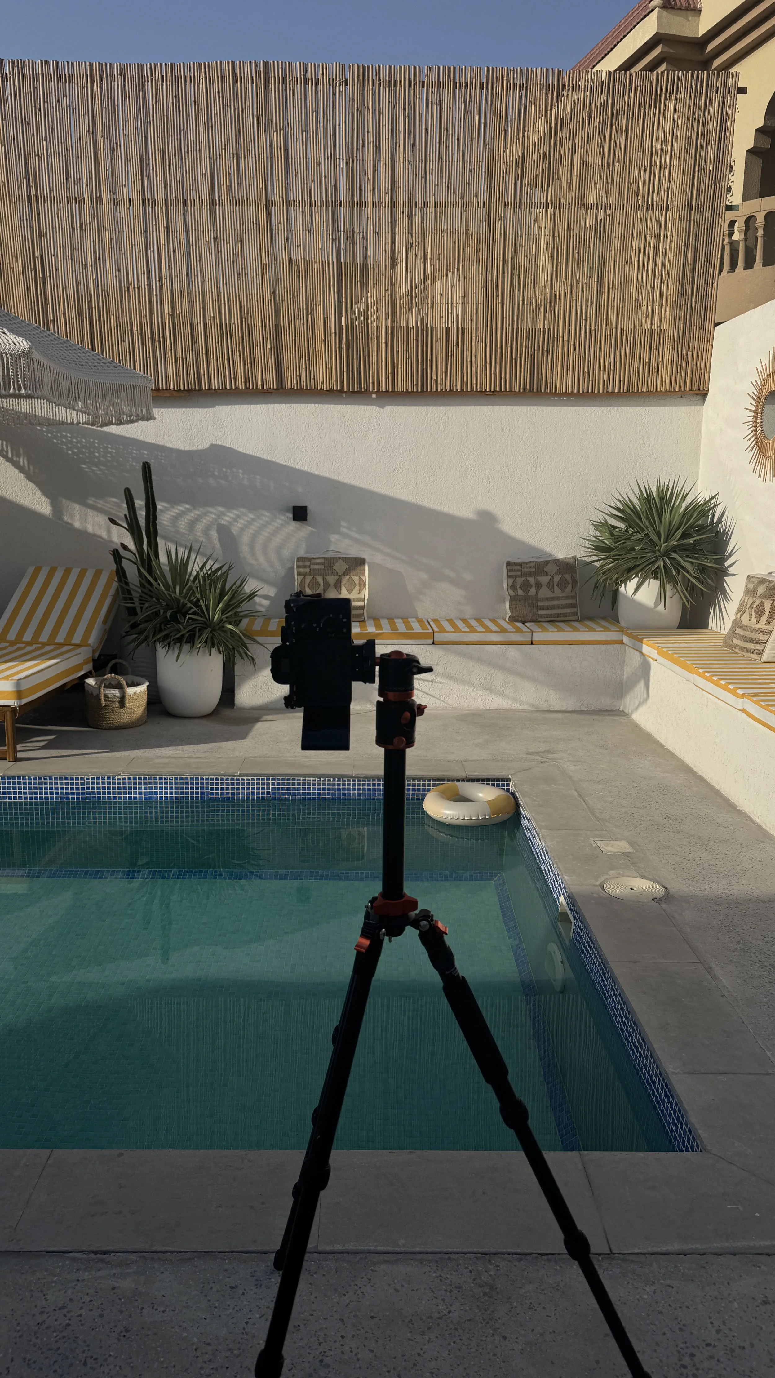 A swimming pool with yellow and white striped lounge chairs and potted plants. A bamboo fence with a number 3 painted on it in the background. A camera on a tripod in the foreground.