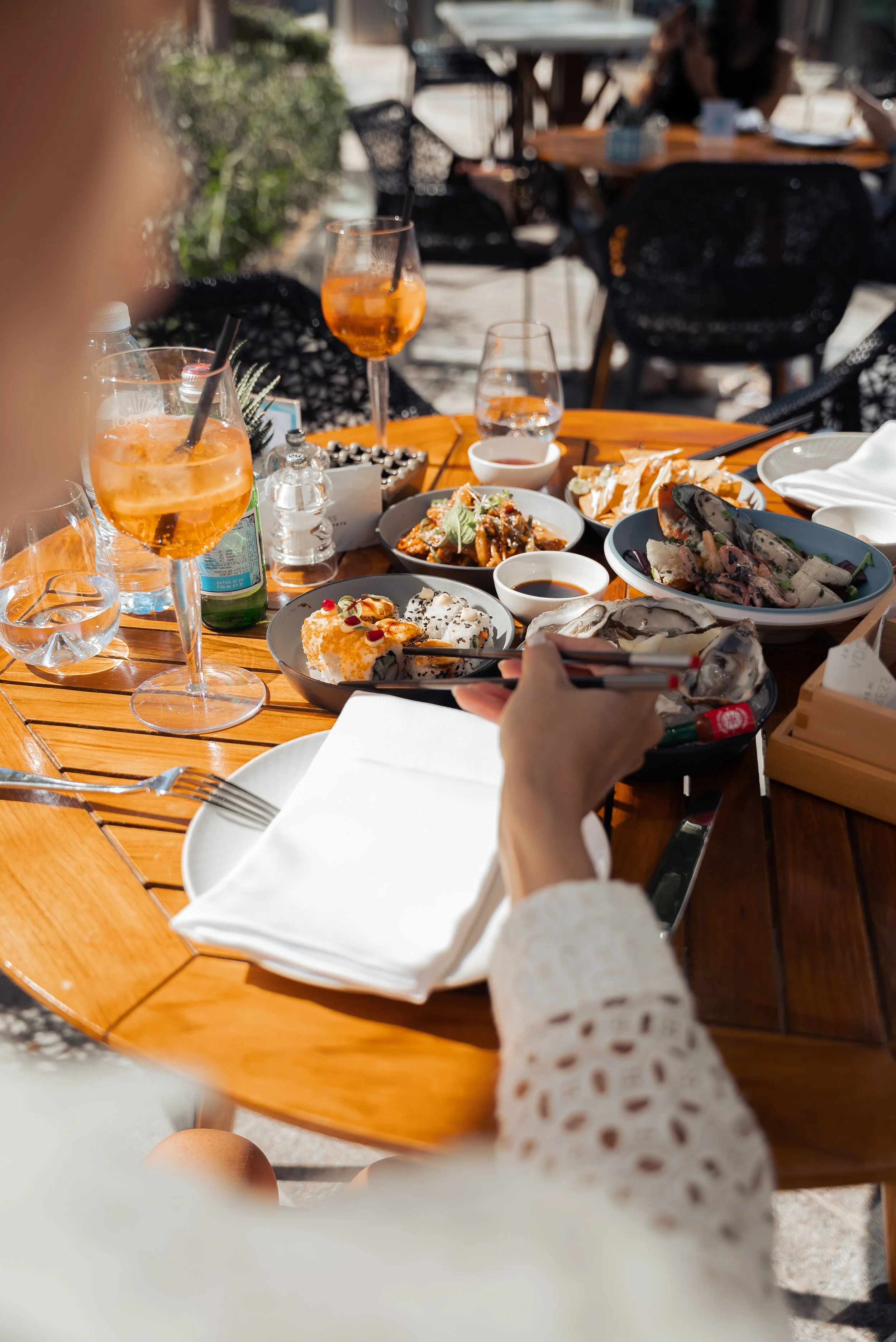 A table set with various dishes and drinks at an outdoor restaurant, with people dining in the background.