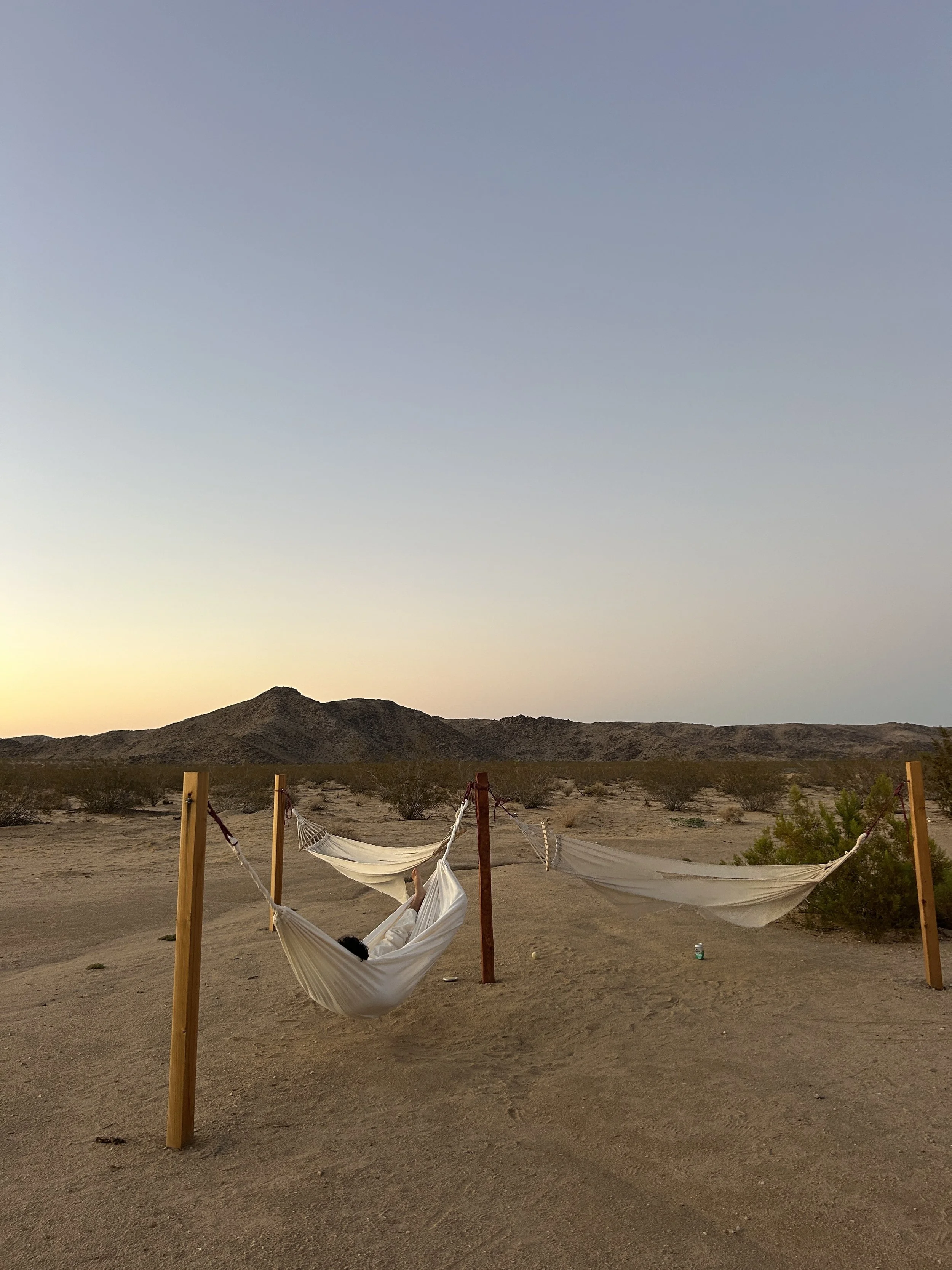 Two hammocks hung between wooden posts in a desert landscape with hills and sparse vegetation at sunset.