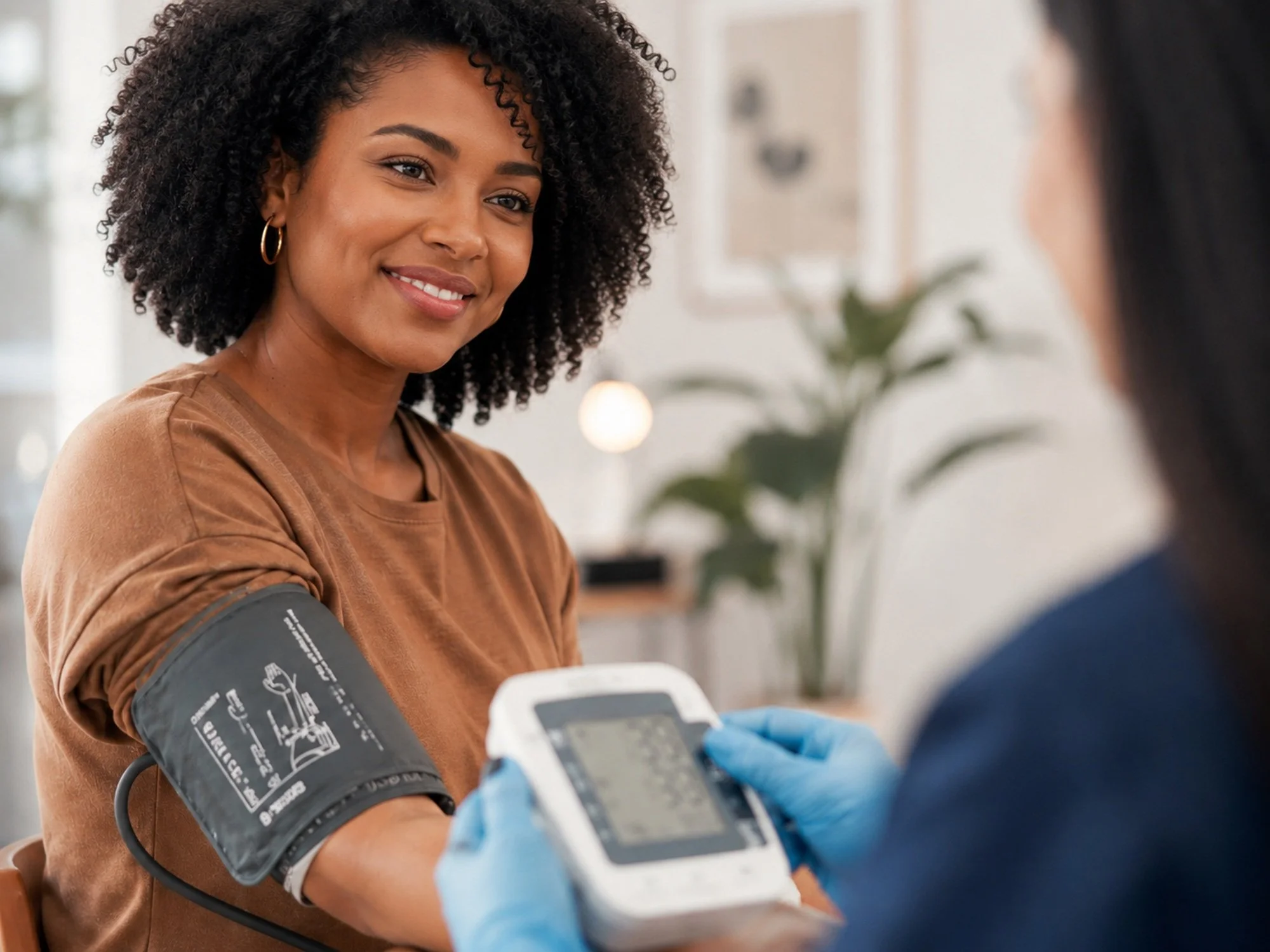 A woman with curly hair smiling as she gets her blood pressure taken by a healthcare professional in a clinical setting.