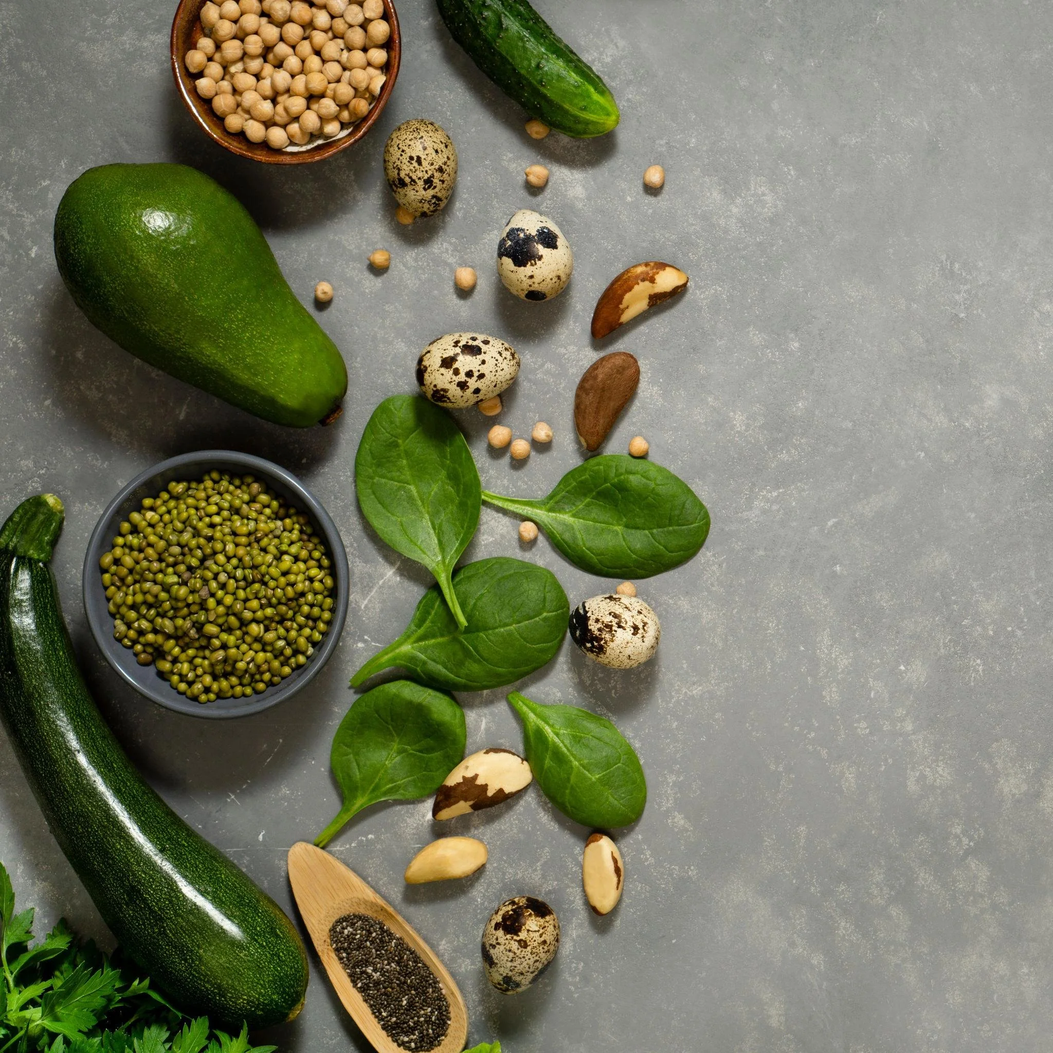 Fresh vegetables and seeds on a gray surface, including an avocado, cucumber, spinach leaves, cracked eggs, and bowls of chickpeas, green mung beans, and black chia seeds.