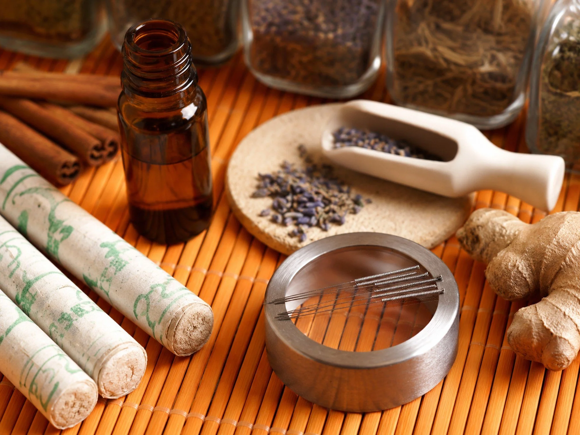 Small amber glass bottle, dried lavender flowers on a beige disc, cinnamon sticks, and ginger root all arranged on a bamboo mat.