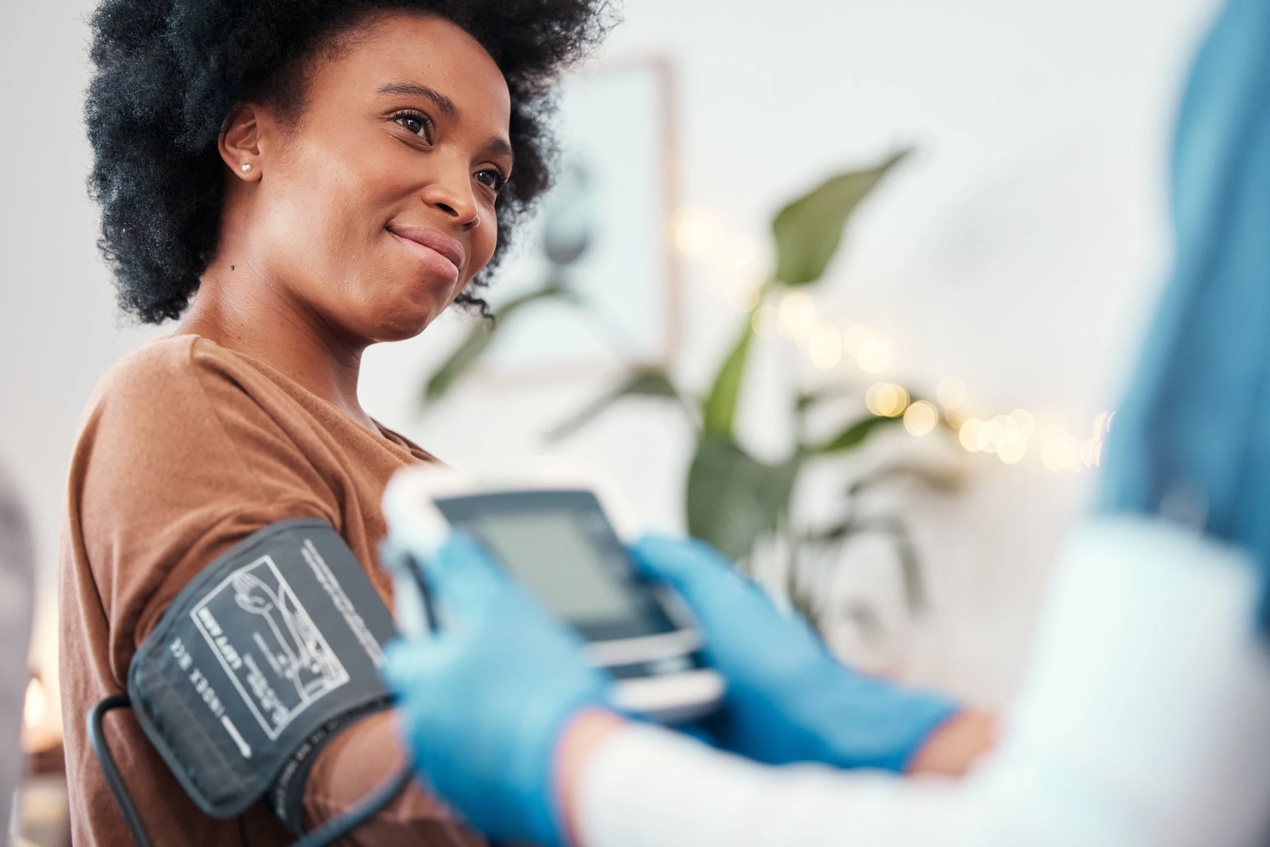 A woman with short curly hair, wearing a brown shirt, receives her blood pressure checked with a cuff on her upper arm, in a bright indoor setting with blurred green plants in the background.
