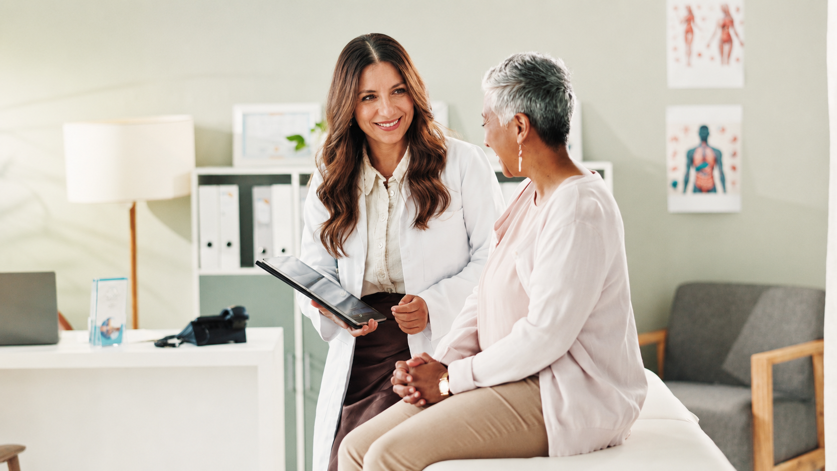 A healthcare professional and a patient having a conversation in a medical office. The professional is holding a tablet and smiling at the patient, who is seated on an examination table.