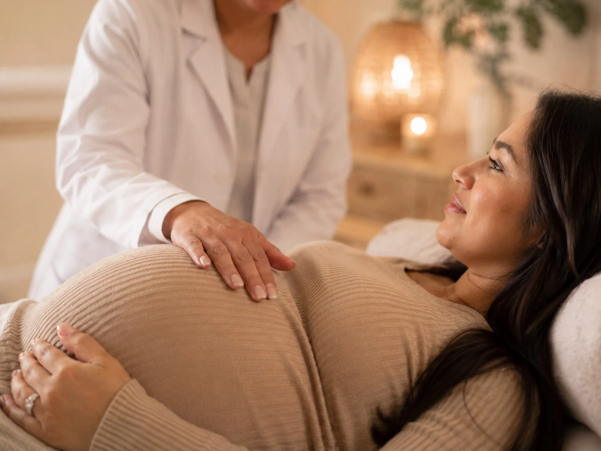 A pregnant woman lying on her side on a bed, smiling, with her hands resting on her belly. A healthcare professional is gently placing a hand on her shoulder, likely during a prenatal checkup. The background includes warm lighting and decorative objects.