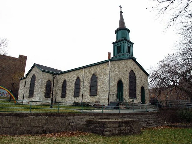 A small stone church with stained glass windows and a steeple, surrounded by a metal railing and a grassy area.