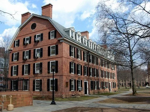 A large, historic brick building with multiple floors and rows of windows, surrounded by trees and a pathway in a park-like setting.