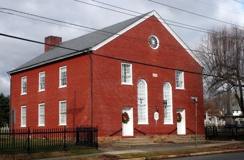 Red brick building with white-framed windows and a gabled roof, decorated with holiday wreaths at the entrance, surrounded by a black metal fence.