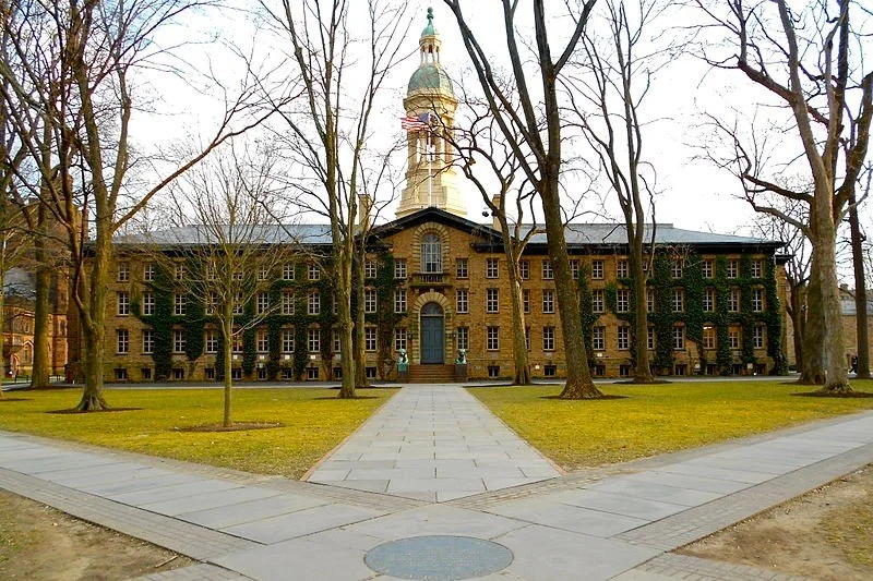Historic brick building with a clock tower, surrounded by leafless trees and a paved walkway leading up to the entrance.