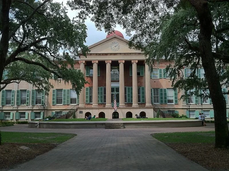 A historic building with a prominent facade featuring large columns and a triangular pediment with a clock. The building has multiple stories with windows and a staircase entrance. There are trees framing the scene and a paved walkway leading to the front entrance. An American flag is displayed in front.