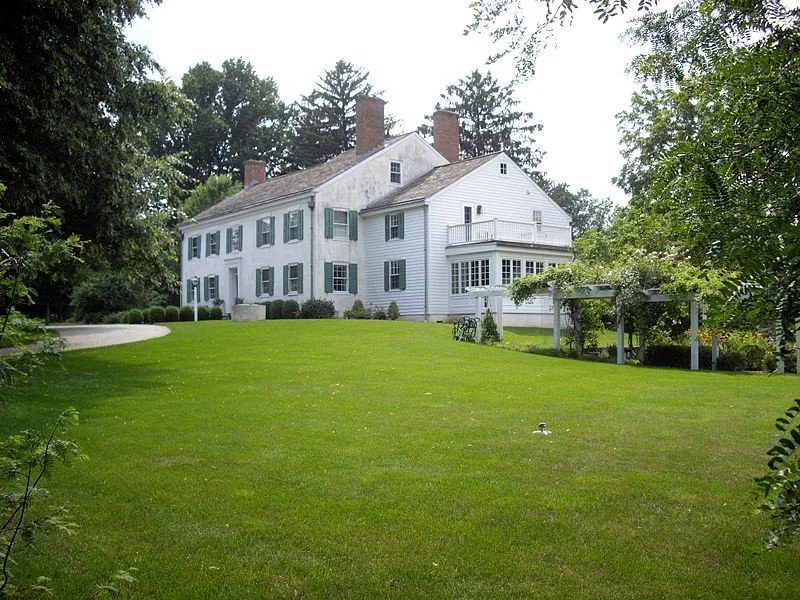A large, white, historic house with multiple chimneys and green shutters, surrounded by a well-maintained lawn and trees.