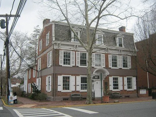 A three-story brick building with white window frames and a white arched entrance, located on a street with a tree in front and power lines to the side.