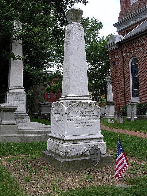 White marble monument with an inscription, American flag nearby, in a cemetery with other tombstones and a brick building in the background.