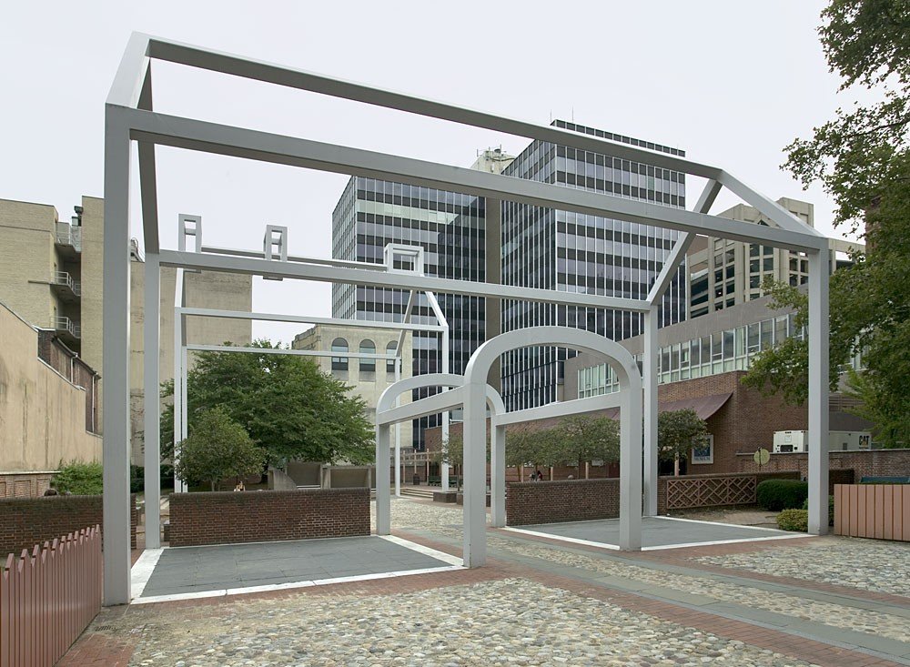 Empty outdoor area with modern white metal arch structures, cobblestone pavement, surrounded by trees and urban buildings.