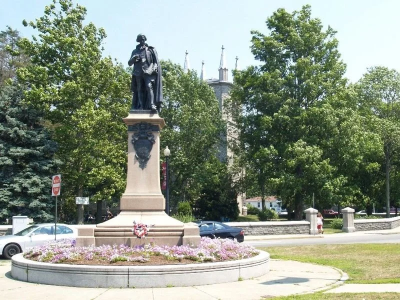 Statue of a historical figure on a tall pedestal surrounded by flowers, with trees and a church in the background.