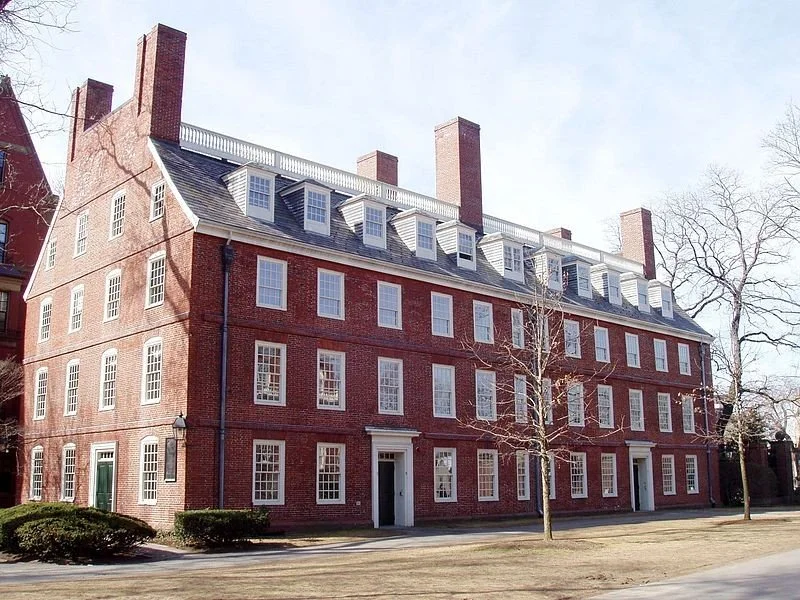 A large red brick building with four floors, white-framed windows, and a steep roof with dormer windows and multiple chimneys. There are leafless trees in front and a grassy area.