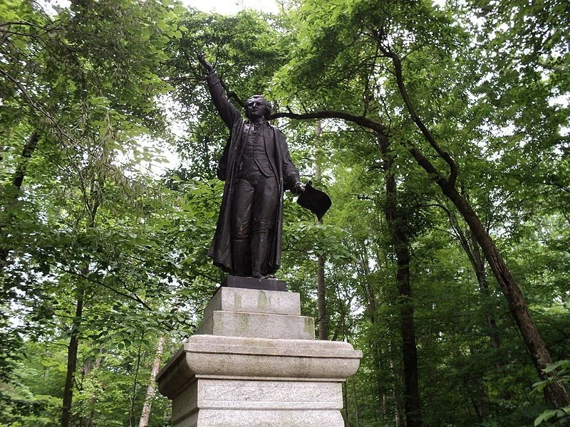 Bronze statue of a man in Victorian attire, holding a hat in one hand and pointing upwards with the other, atop a stone pedestal in a wooded area.