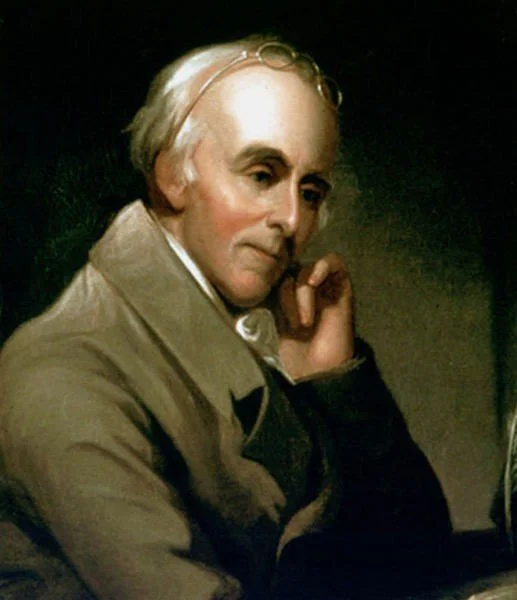 A portrait of a man with gray hair, wearing a beige coat, resting his face on his hand, with a contemplative expression.