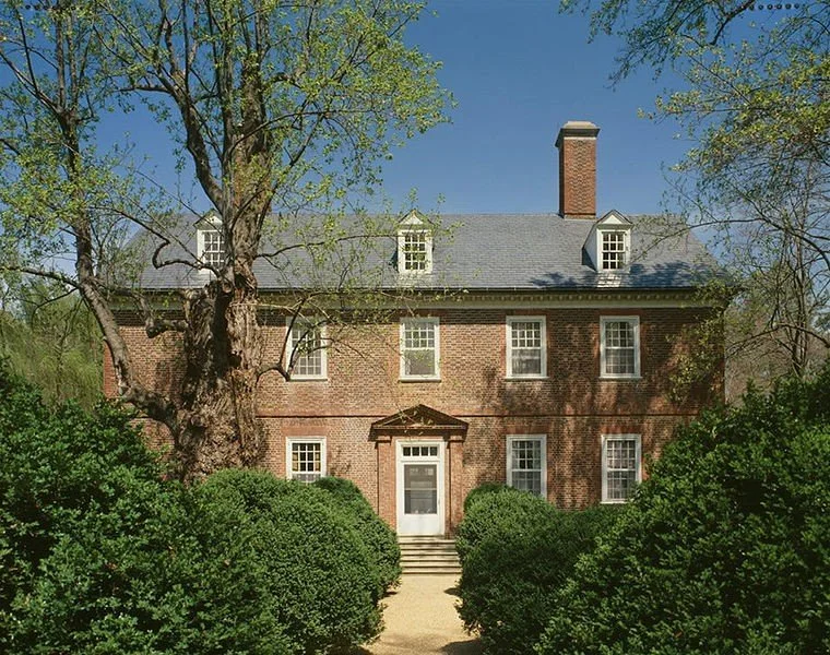 A two-story brick house with a gabled roof, white window frames, a central front door, and surrounded by green bushes and trees under a blue sky.
