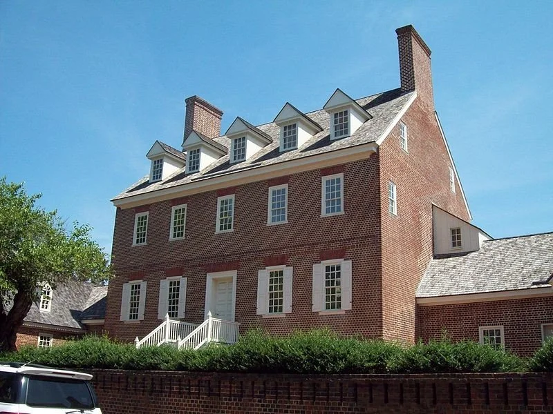 A large brick house with multiple windows and dormer windows on the roof, surrounded by greenery and trees, under a clear blue sky.