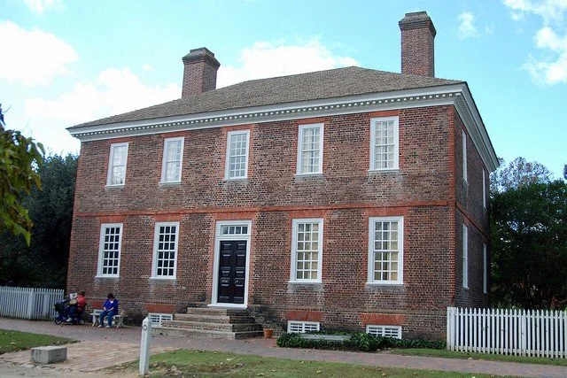A large two-story brick house with white-framed windows and a black front door, with steps leading up to the entrance, surrounded by a white picket fence and some trees.