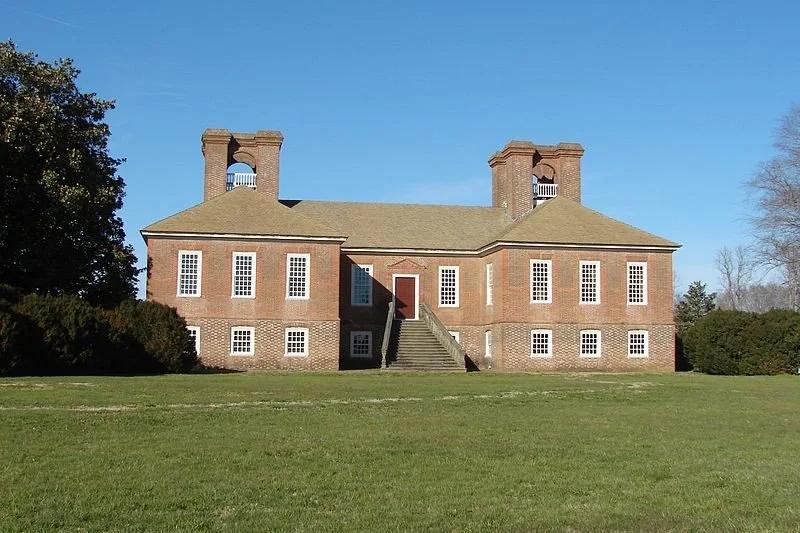 A historic brick building with two prominent chimneys, small windows, and stairs leading to a central door, set against a clear blue sky with green grass in front.