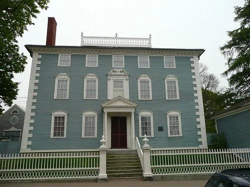 A large, light blue historic house with white trim, three stories, a staircase leading to the front door, and a white picket fence.