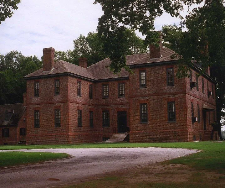Large red brick historic mansion with multiple chimneys, surrounded by green trees and a gravel driveway in front.