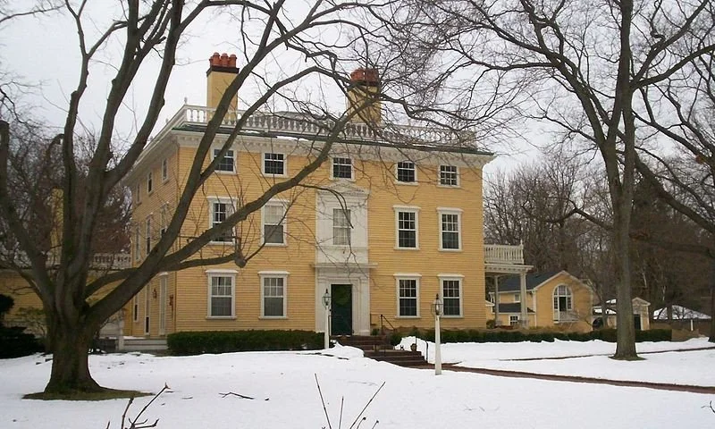 A large yellow three-story house with white trim, brick chimneys, and a balcony, surrounded by leafless trees and snow on the ground in winter.