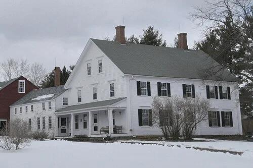 A large white historic house with black shutters, a covered porch, and snow on the ground, set in winter.