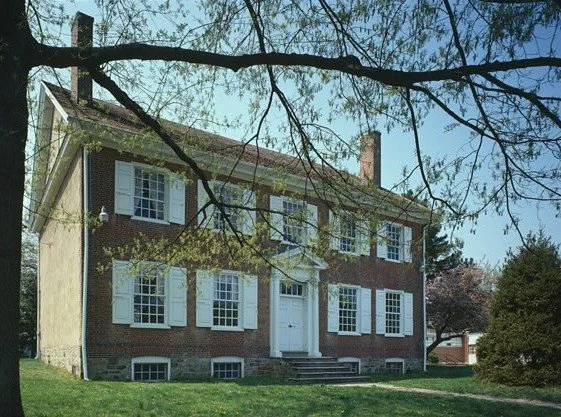 A two-story brick house with white window shutters and a white front door, surrounded by a grassy lawn and trees.