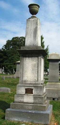Gravestone with a large urn on top, located in a cemetery with other tombstones and trees in the background.