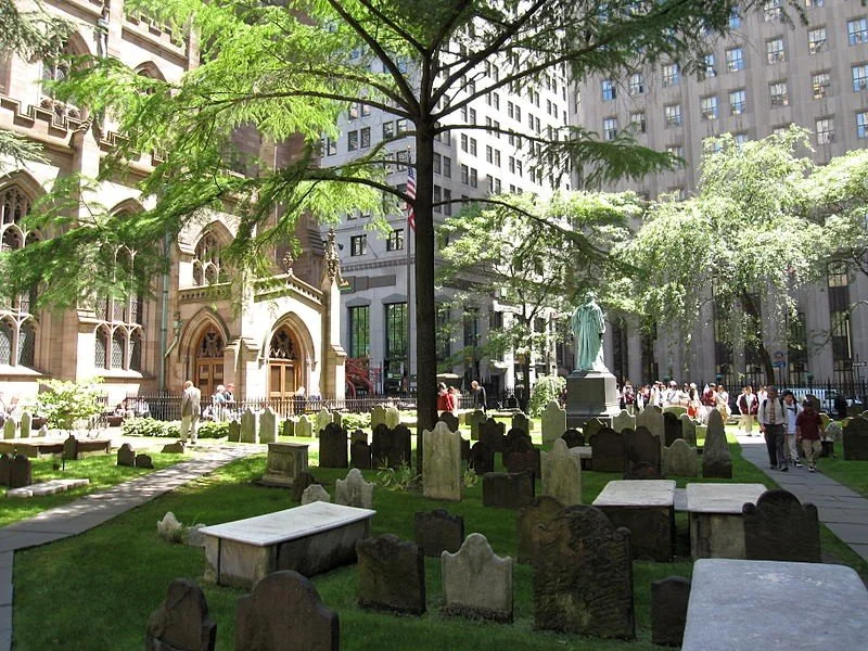 City park with visible tombstones and a statue, surrounded by tall buildings and people walking.