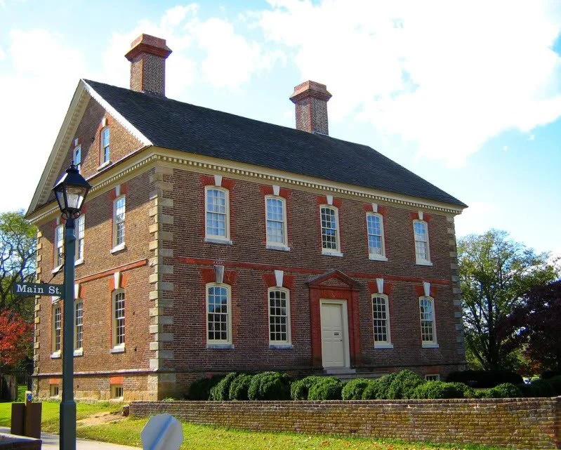 A three-story historic brick building with white window trims, a centered white door, and two brick chimneys, located at the corner of Main Street. The building is surrounded by a low brick wall and trimmed bushes, with a street lamp and a street sign visible.