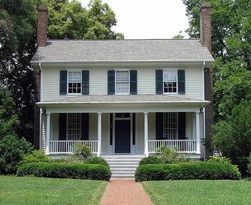 A two-story white house with black shutters, a front porch, and brick chimneys on each side, surrounded by green shrubs and trees.