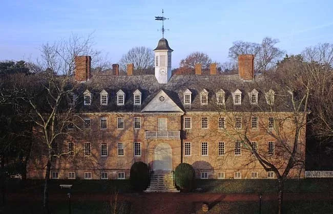 A large historic brick building with a central tower, multiple chimneys, and a symmetrical facade surrounded by trees.