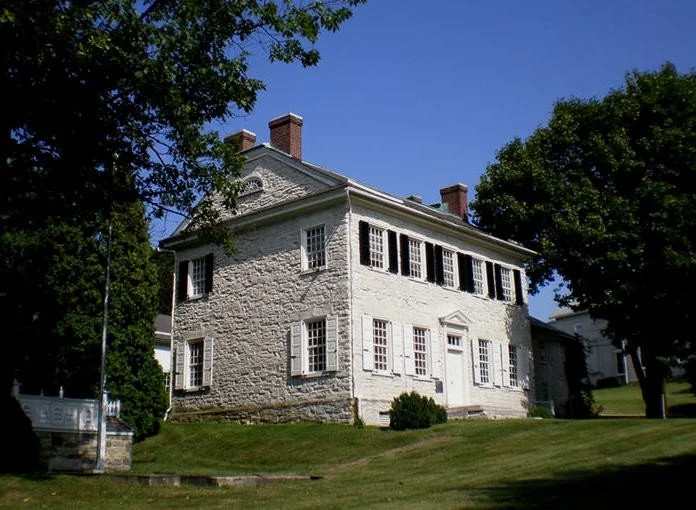 White, two-story historic house with multiple windows, located on a grassy hill with trees and a clear blue sky.