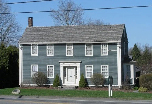 A classic two-story house with blue siding, white trim, and multiple windows. It has a brick chimney, a small front porch, and is surrounded by a grassy yard with bushes.