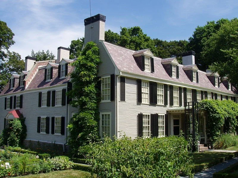 Large historic white house with black shutters, a steep metal roof, multiple chimneys, surrounded by trees and a garden.