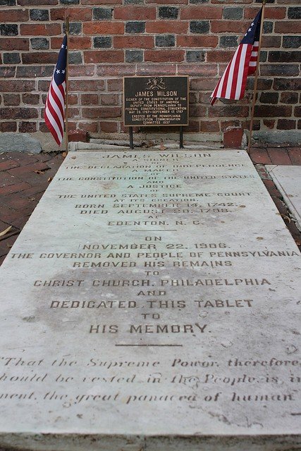 A memorial plaque dedicated to James Wilson, located in Philadelphia. It includes American flags on either side and a brick wall behind. The plaque commemorates his life and service.