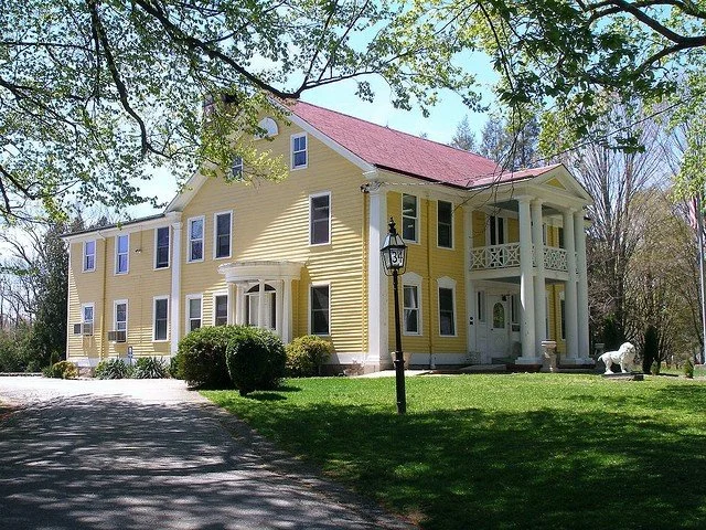 Yellow multi-story house with white trim and a red roof, surrounded by trees and greenery.