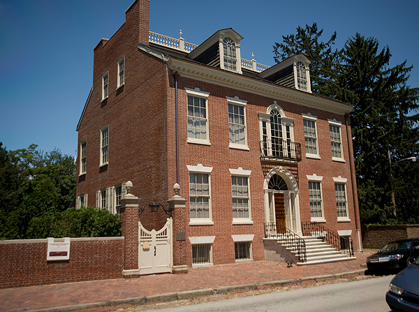 A large three-story brick house with white trim, multiple windows, and a small front porch with steps leading up to the door. The house is surrounded by trees and a brick wall.