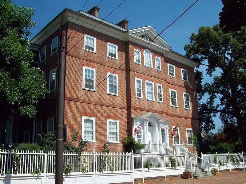A large three-story brick building with white window frames, white decorative trim, and a white porch with steps. The building has an American flag hanging near the entrance, and is surrounded by a white picket fence and greenery. Tall trees are visible around it under a clear blue sky.