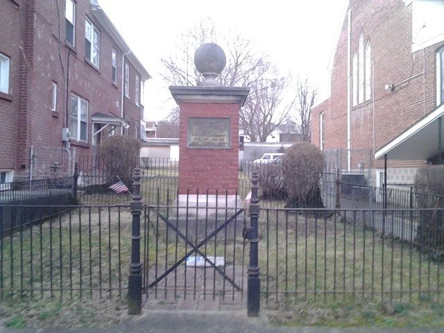 Small fenced park area with a brick monument or memorial in the center, topped by a black sphere and surrounded by houses and trees.