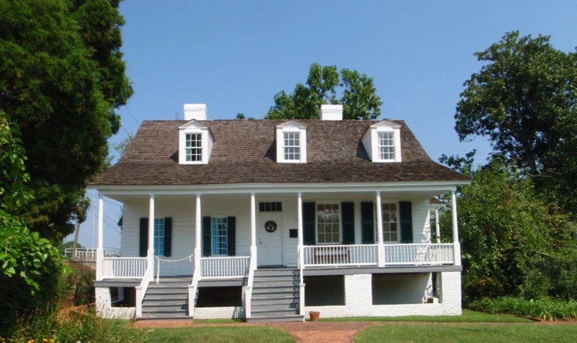 A white two-story house with dormer windows, a front porch with bunk doors, and surrounded by green trees and grass.