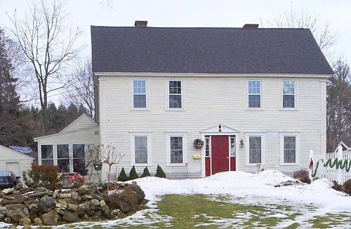 A large white two-story house with a red front door, snow on the ground, and a yard with rocks and trees in the background.