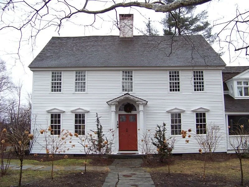 White two-story house with a red front door, symmetrical windows, and a small front porch surrounded by leafless bushes and trees.