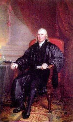 Painting of a man with white hair wearing black robes, sitting in a red armchair in a room with a patterned rug and draped curtain.
